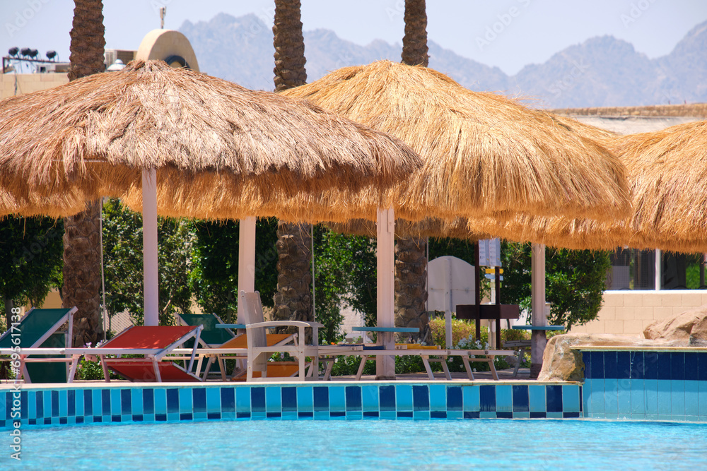 Empty deck chairs under straw shade umbrellas on swimming pool side in tropical resort. Summer vacations and getaway concept Empty deck chairs under straw shade umbrellas on swimming pool side in tropical resort. Summer vacations and getaway concept