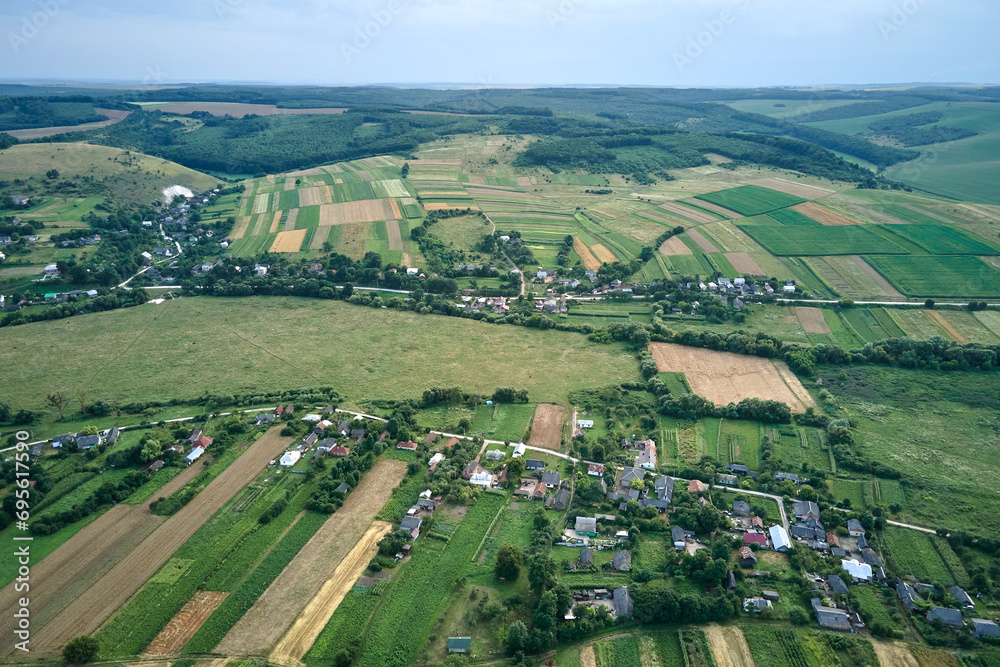 Aerial landscape view of village houses and distant green cultivated agricultural fields with growing crops on bright summer day