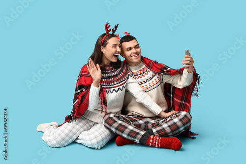 Happy young couple in Christmas pajamas taking selfie on blue background