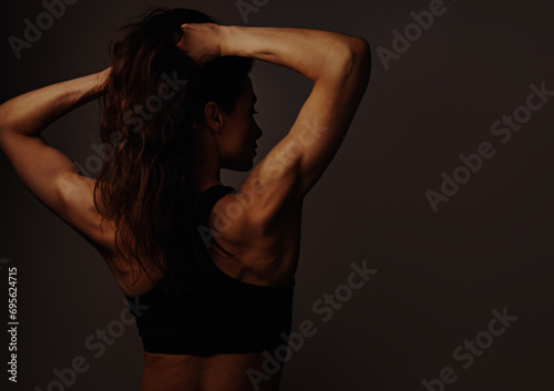 Sport athletic muscular woman with long curly hair doing stretching workout for shoulders, blades and arms in sport black bra, standing on dark shadow background with empty space. Back view.