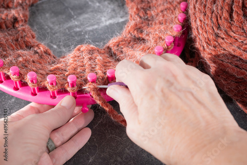 Soft, wool, yarn in a burnt orange color.  Hands using a hook tool on a pink, round knitting loom with both knit and pearl stitches. photo