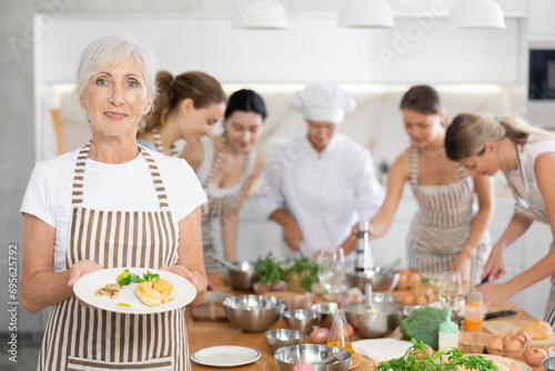 Senior lady visiting culinary school holds plate with cooked chicken fillet in hands, successful result of studies