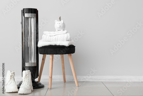 Electric heater and piggy bank with clothes on stool near white wall. Heating season concept photo