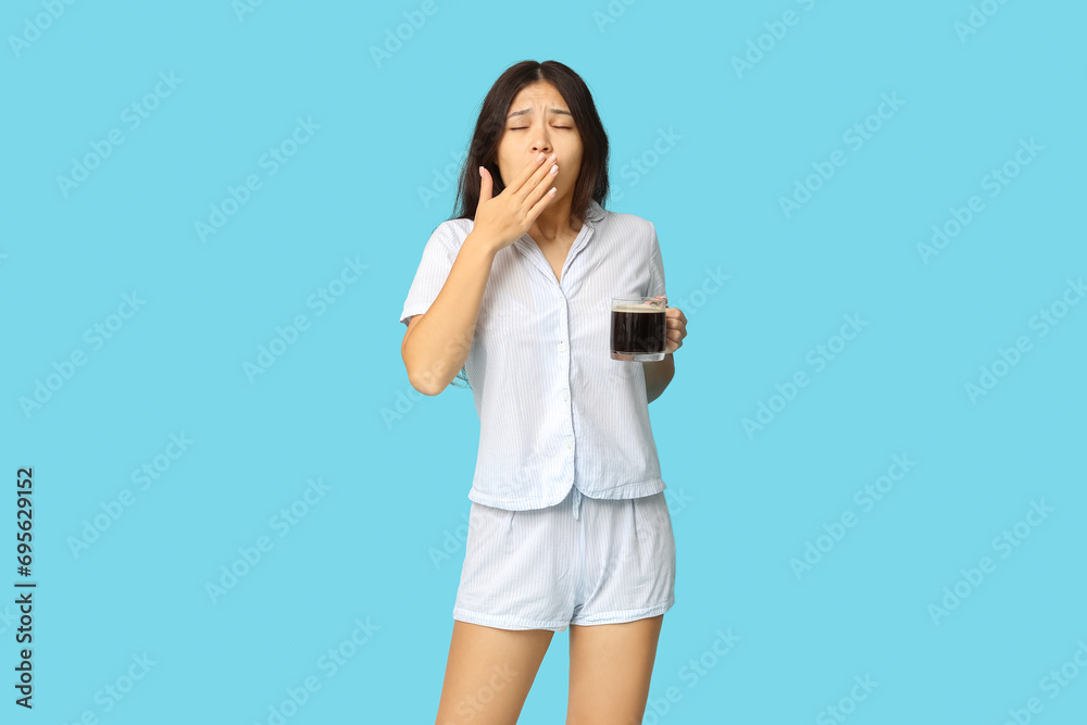 Beautiful young Asian woman in pajamas with cup of coffee yawning on blue background Beautiful young Asian woman in pajamas with cup of coffee yawning on blue background