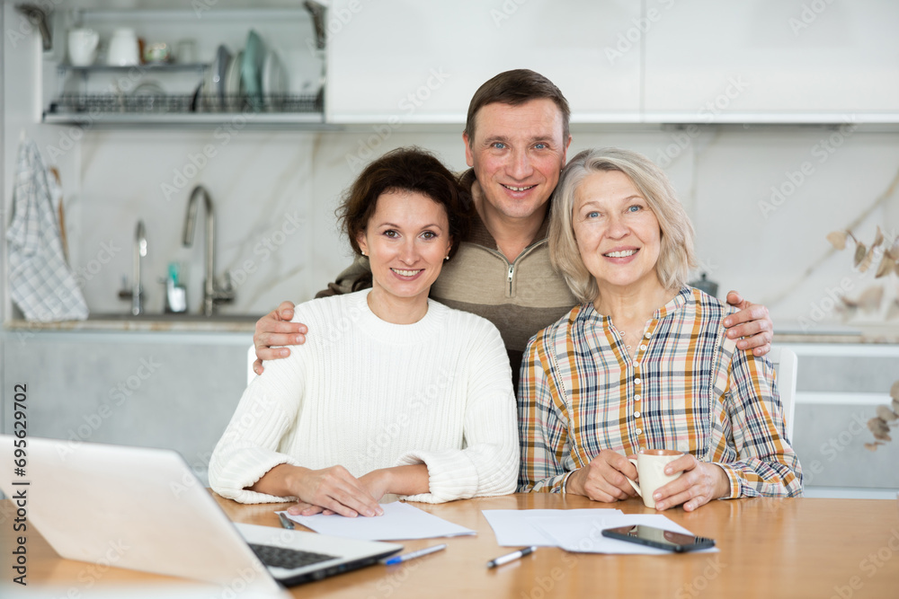 Obraz premium Smiling middle-aged man and two women posing at the kitchen-table with sheets of paper and laptop in front of them