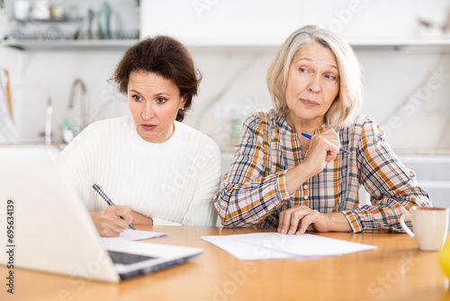 Attentive old and middle-aged women joining distant education using laptop in the kitchen