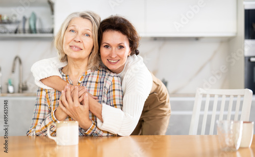 Smiling old woman sitting at the kitchen table and middle-aged woman embracing her kindly