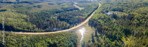 Aerial panoramic view of a lush norther forest with a curving gravel road and the sun reflecting off the water in a small pond. 