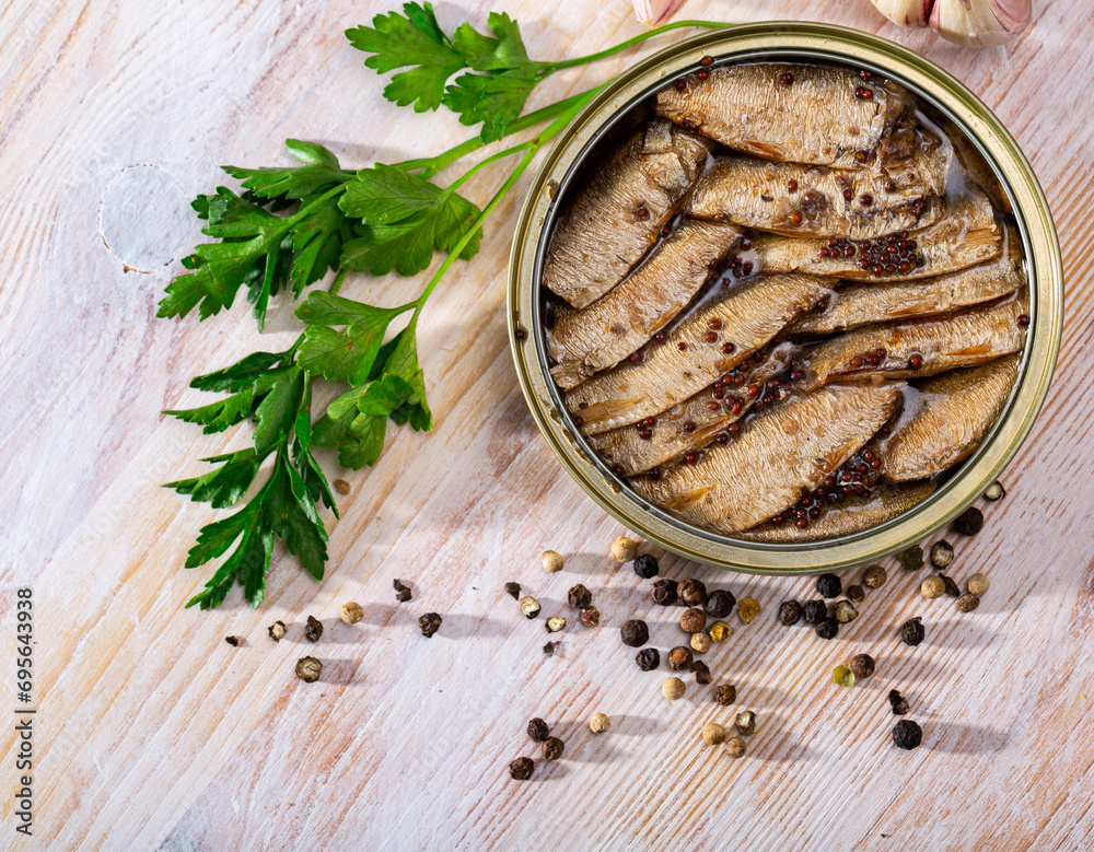 Open can of spicy smoked sprats in oil on wooden table with peppercorn, garlic and parsley sprig..