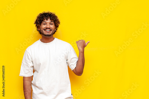 young indian curly guy pointing at copy space on yellow isolated background and smiling