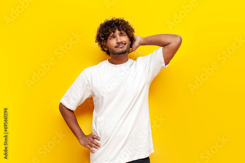 young indian puzzled guy thinks and remembers and scratches his head on yellow isolated background photo