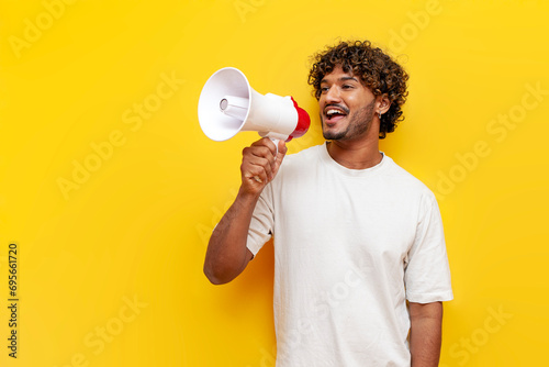 young indian guy announces information into a megaphone on a yellow isolated background photo