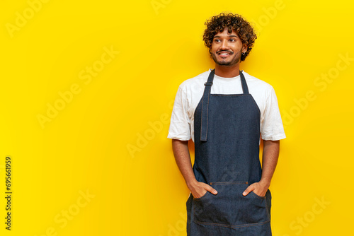 young indian guy barista in an apron stands and looking at copy space on yellow isolated background