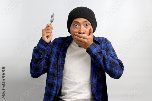 Surprised young Asian man, clad in a beanie hat and casual shirt, covers his mouth in shock and embarrassment, displaying a shocked expression after making a mistake, while holding a fork photo