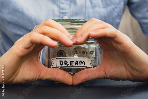 Unrecognizable woman showing heart sign Saving Money In Glass Jar filled with Dollars banknotes. DREAM transcription in front of jar. Managing personal finances extra income for future insecurity