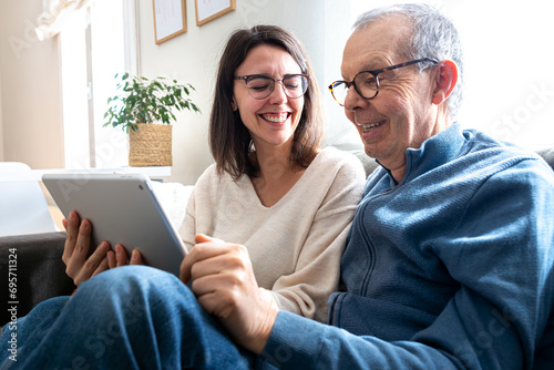 Happy senior man and his young daughter laughing together using digital tablet sitting on sofa at home.