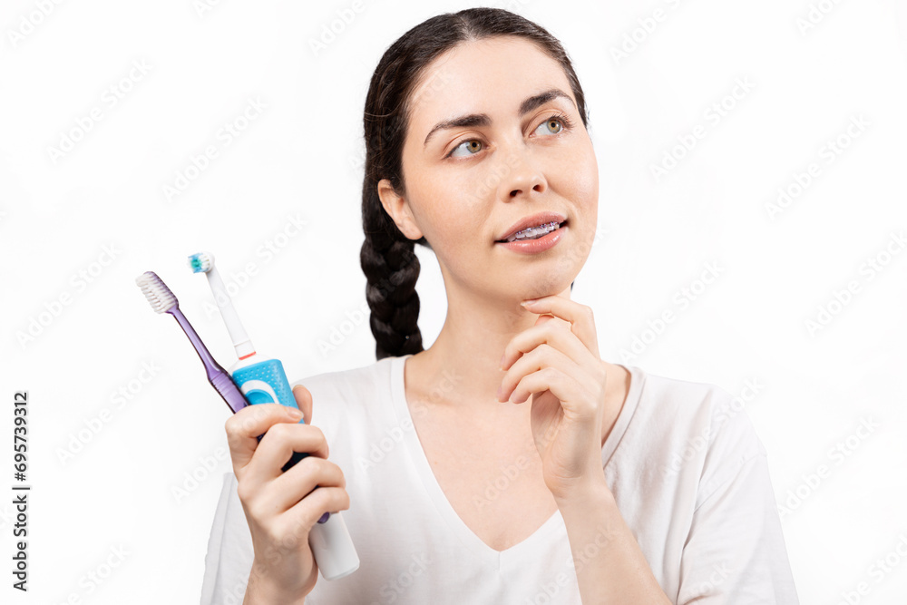 Portrait of young Caucasian woman with ligature braces on her teeth, thinking about choose between electric and manual toothbrush. White background. Concept of orthodontic treatment and dental care Portrait of young Caucasian woman with ligature braces on her teeth, thinking about choose between electric and manual toothbrush. White background. Concept of orthodontic treatment and dental care