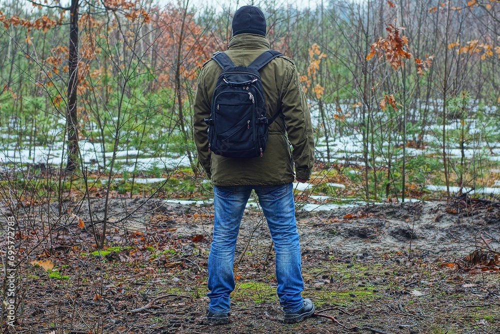 Naklejka premium fashionable black synthetic stylish with plastic zippers closed vintage backpack worn on the shoulders of a man dressed in military olive green a stylish jacket and a knitted black hat in the forest