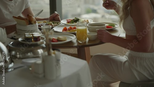 A cinematic shot with unrecognizable faces. A young couple, a man and a woman in white clothes, are sitting having breakfast in a hotel on the top floor of a skyscraper. Healthy meals at the hotel photo