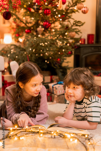 boy and girl have fun near Christmas tree, lay on floor reading a book near flashing lights, smiling and laughing during holidays