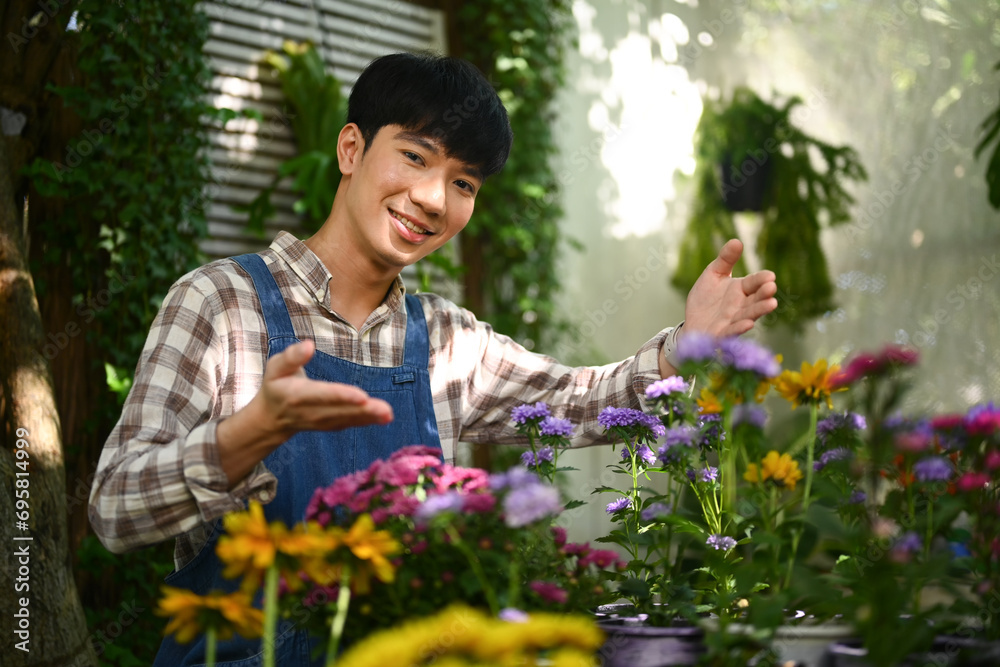 Smiling Asian male florist wearing apron standing among flowers at his shop.