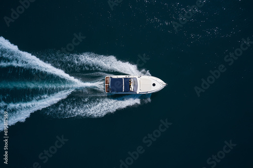 A white boat with a blue awning moves quickly on dark blue water, leaving white traces, top view.
