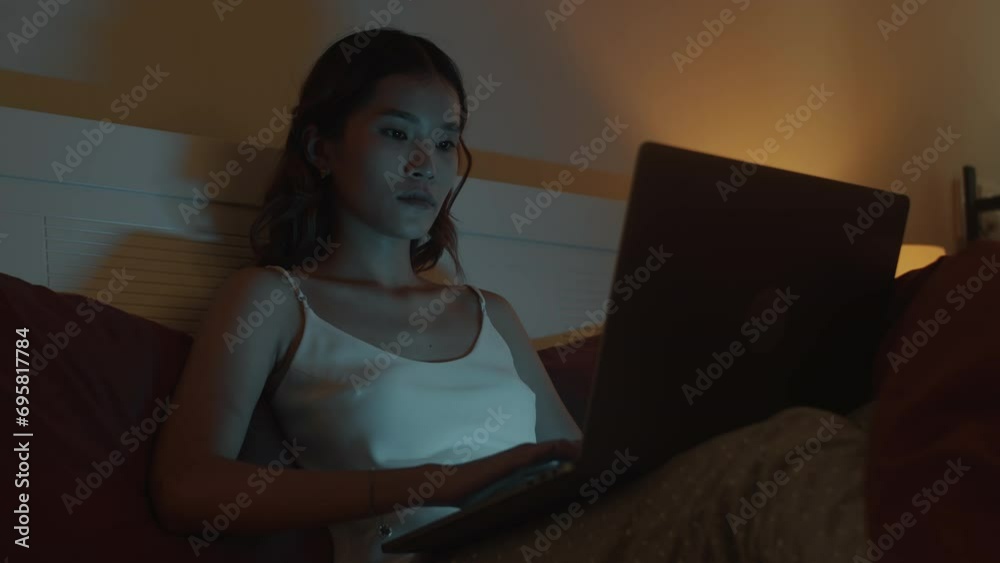 Medium shot of serious woman in homewear working on laptop late at night while sitting on bed