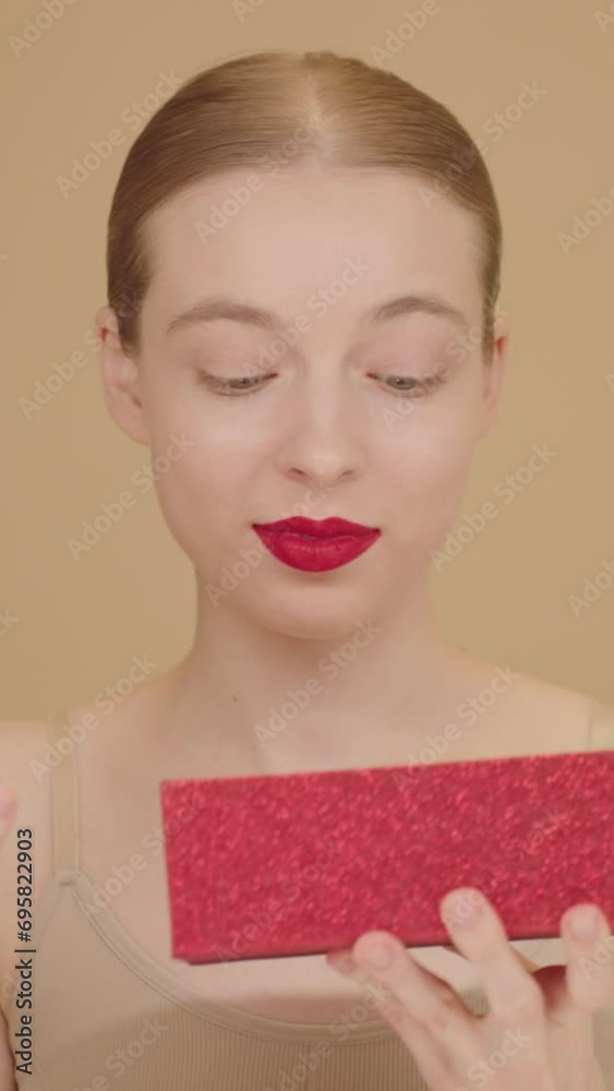 Zoom Out Effect. Portrait of a delightful woman with red lips opening a red gift box and looking inside with a surprised expression. Excited young woman receiving gift. Studio shot on beige background Zoom Out Effect. Portrait of a delightful woman with red lips opening a red gift box and looking inside with a surprised expression. Excited young woman receiving gift. Studio shot on beige background
