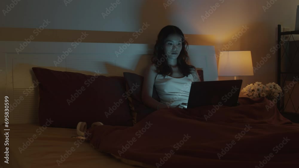 Medium long shot of pensive female workaholic doing report on wireless computer while sitting in bed at nighttime Medium long shot of pensive female workaholic doing report on wireless computer while sitting in bed at nighttime