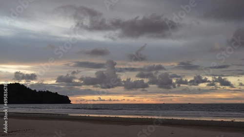 Twilight after sunset on a tropical island. Purple cumulus clouds in the sky, illuminated by orange light above the horizon. Ocean waves, foaming, roll onto a sandy beach. A hill in the distance.