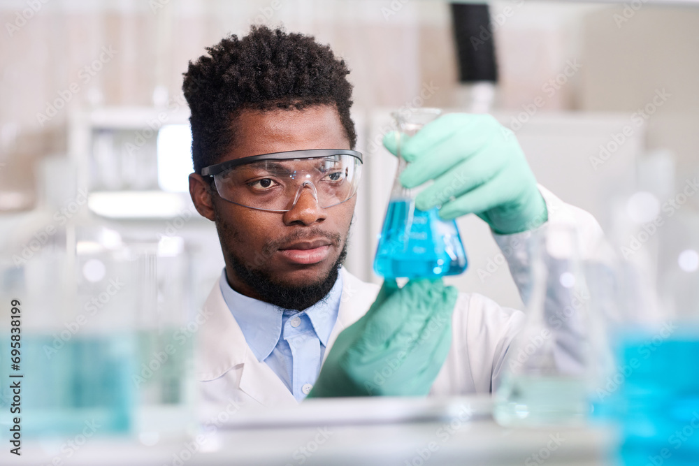 Close up portrait of young African American male laboratory worker in gloves and protective glasses looking at conical flask with blue liquid substance Close up portrait of young African American male laboratory worker in gloves and protective glasses looking at conical flask with blue liquid substance