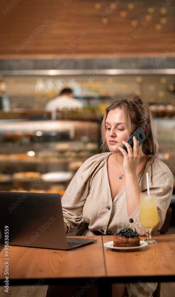 Young freelancer working remotely online sitting with laptop and speaking with smile on phone with her superior in cafe indoor interior Young freelancer working remotely online sitting with laptop and speaking with smile on phone with her superior in cafe indoor interior