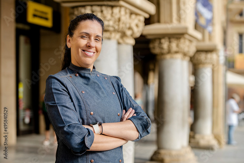 Woman chef wears blue coat, poses with arms crossed and broadly smiles outdoors on the square. Restaurant worker, culinary gourmet, pastry chef.
