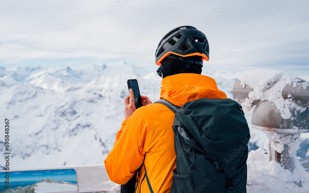 Man with orange jacket in winter Alps. Man Doing Photography With Mobile Phone Over Snowy Landscape