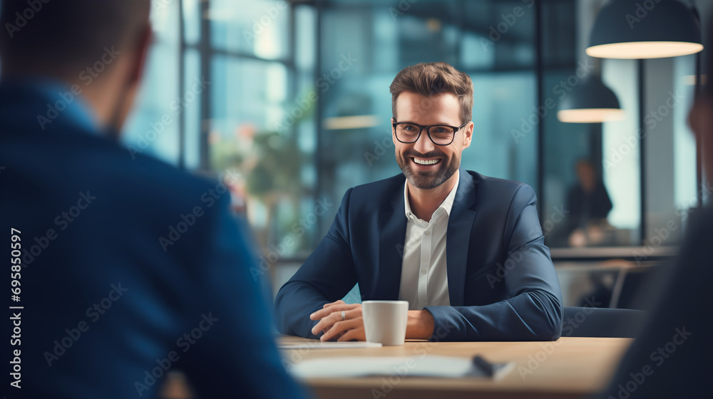 Handsome young businessman wearing an elegant suit with glasses, having a meeting with company employees or workers, sitting at the office table, corporation future plans, job hiring interview - obrazy, fototapety, plakaty 