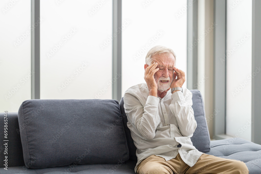 Man presses a hand to head and suffers from unbearable pain, Senior man having a headache and touching his head while suffering from a migraine in the living room Man presses a hand to head and suffers from unbearable pain, Senior man having a headache and touching his head while suffering from a migraine in the living room