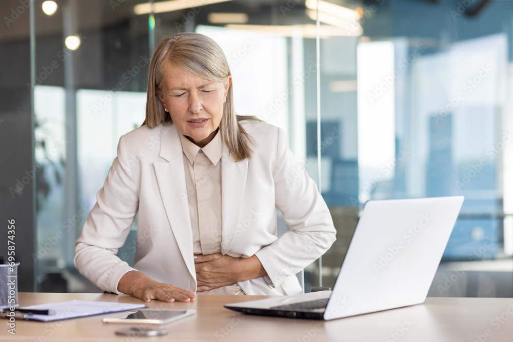 Sick overworked woman at workplace inside office has severe stomach pain, senior mature business woman holding hands on stomach massaging while sitting at desk with laptop. Sick overworked woman at workplace inside office has severe stomach pain, senior mature business woman holding hands on stomach massaging while sitting at desk with laptop.