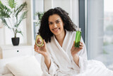 Beautiful calm female in bathrobe looking at camera with avocado half in hand while holding cosmetic oil bottle in bedroom interior. Young woman appreciating green fruit for great skin benefits.