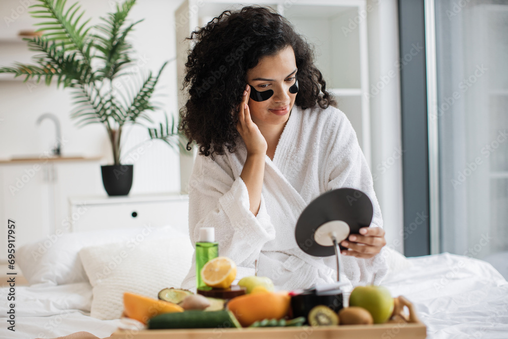 Pretty lady keeping morning skincare routine in bedroom. Smiling African female applying under-eye black masks after morning bath while looking at makeup mirror on bed tray with home spa essentials. Pretty lady keeping morning skincare routine in bedroom. Smiling African female applying under-eye black masks after morning bath while looking at makeup mirror on bed tray with home spa essentials.
