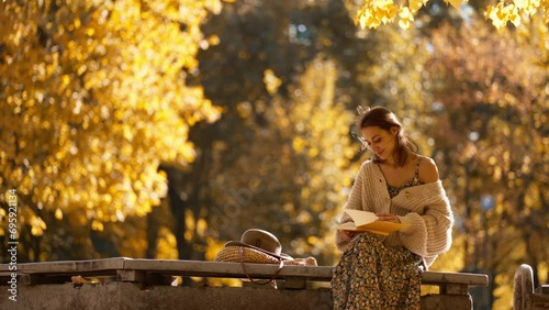 young brunette woman in autumn park with yellow foliage against bright sun with sunflare. stylish attractive girl in dress sitting in park at warm fall day with yellow notebook and writing photo