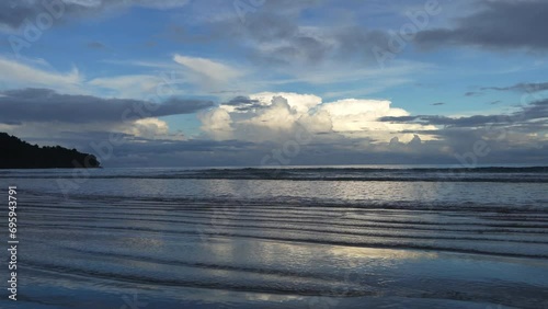 Early morning by the ocean. The clouds in the sky are highlighted in golden. The waves roll towards the shore, spreading over the wet beach. Reflection on the shiny wavy sand. Malaysia. Borneo.