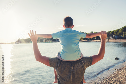Cute little Caucasian boy from behind cheering with arms outstretched while sitting on his father's shoulders at the beach. Playful young child from the back having fun and bonding with dad outdoors. photo
