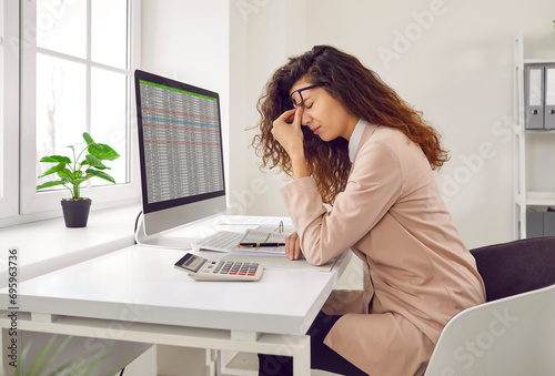 Stressed, tired young woman financial accountant with eye strain and headache holding her nose bridge while sitting at working desk with computer in office. Pain, stress at work, overworking concepts