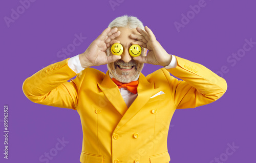 Portrait of funny optimistic senior man with happy emoji eyes. Studio shot of old guy wearing bright yellow jacket and orange bow tie covering his eyes with two smiley faces he's holding in his hands