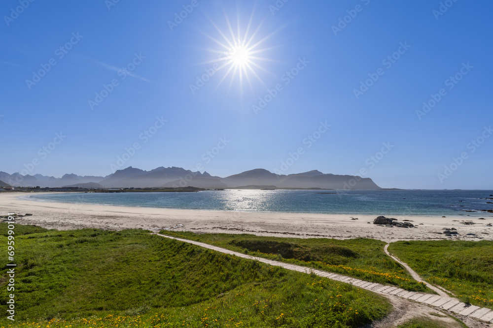 Gleaming sun over Jusnesvika Bay with path cutting through green grass, leading to the white sands of Rambergstranda beach, against a backdrop of distant mountains in Lofoten. Norway
