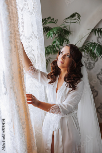 A happy bride is preparing for her luxurious wedding in a hotel room, with a wedding dress on a mannequin nearby. Portrait of a woman with fashionable hair, makeup and a smile in a dressing gown. © Vasil