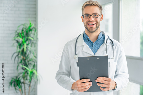 Handsome smiling medical doctor in white coat with clipboard, looking at camera and smiling while standing in clinic photo