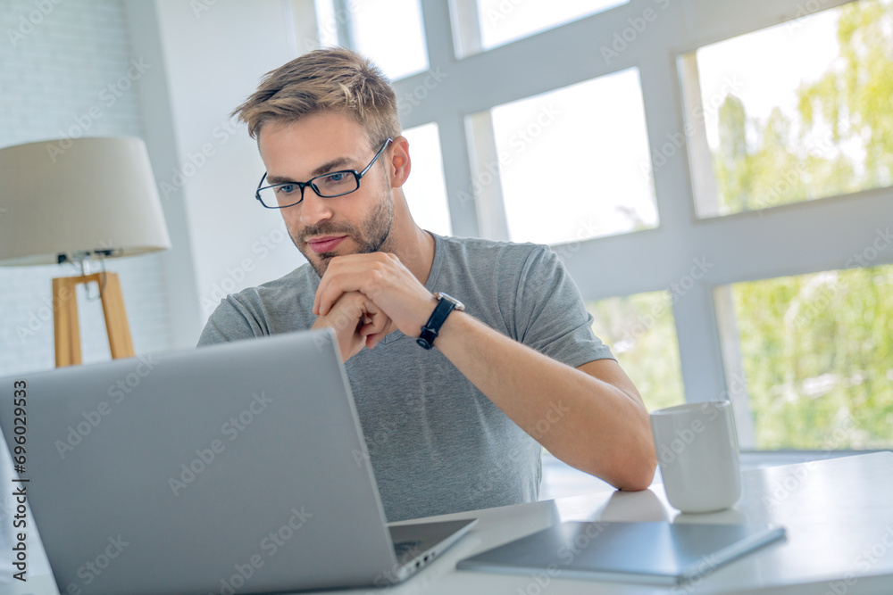 Concentrated serious handsome man in eyeglasses using laptop computer while sitting by the table at office Concentrated serious handsome man in eyeglasses using laptop computer while sitting by the table at office