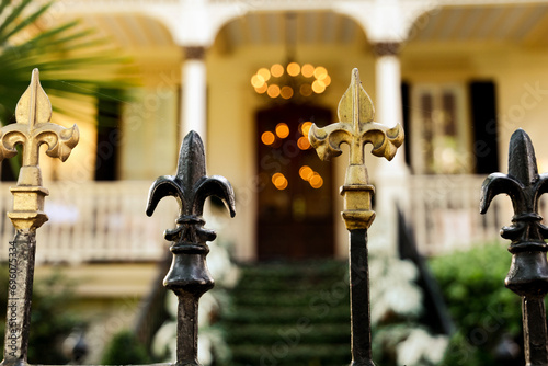 A cast wrought iron fence lined with black and gold fleur de lis post toppers with a New Orleans southern style home in the background photo