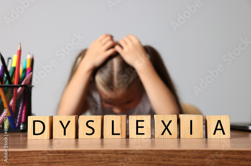 Little girl covering head with hands and cubes with word Dyslexia at wooden table, selective focus photo